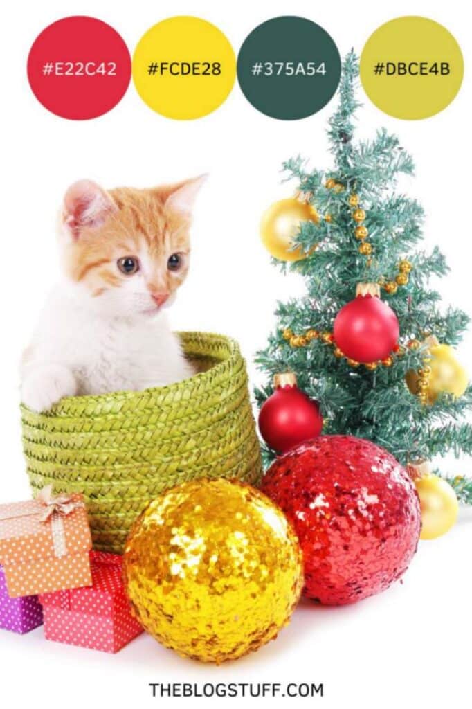 A cute kitten in a green basket surrounded by colorful gift boxes, oversized gold and red baubles, and a small Christmas tree with ornaments.