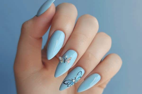 Light blue stiletto nails with 3D butterfly and gem decorations on the ring finger