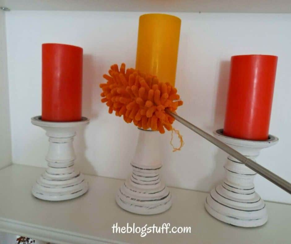 An orange duster cleaning a yellow candle, flanked by two red candles on white holders.