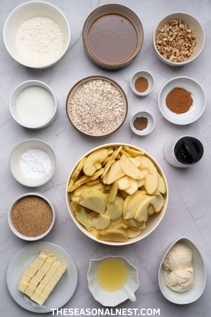 Flat lay of sliced apples, oats, sugar, flour, butter, nuts, spices, and caramel sauce arranged in bowls on a marble surface.