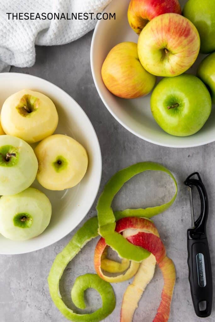 Fresh apples in a bowl, some peeled and cored, with apple peels and a peeler on the counter.