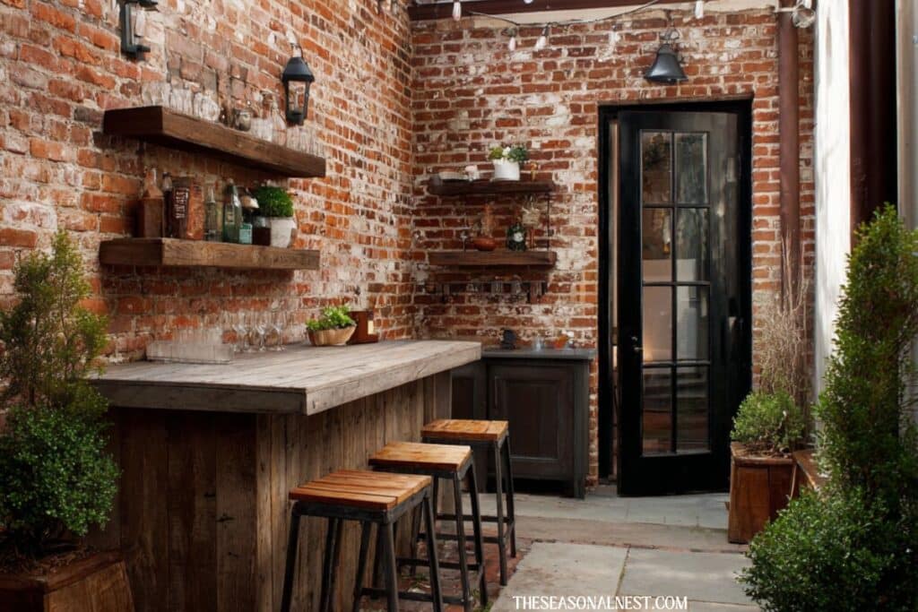 Small rustic backyard bar with built in wooden counter, brick wall, floating shelves, and stools in a narrow patio space.