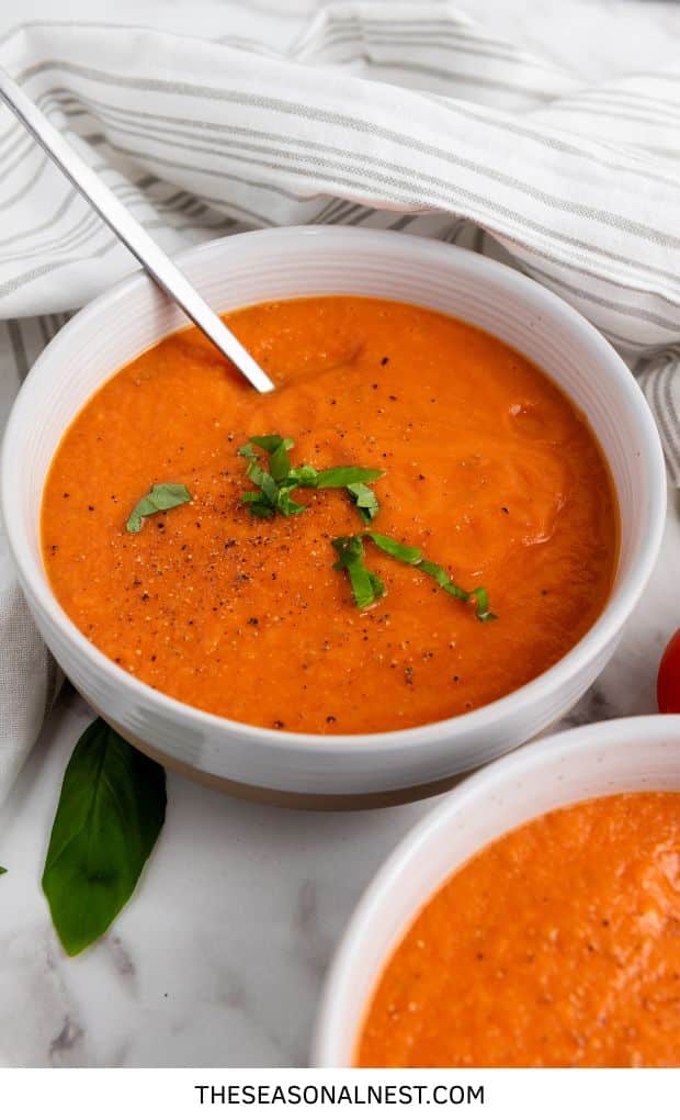 Bowl of roasted tomato basil soup garnished with fresh basil and cracked black pepper, served with a spoon on a marble surface.