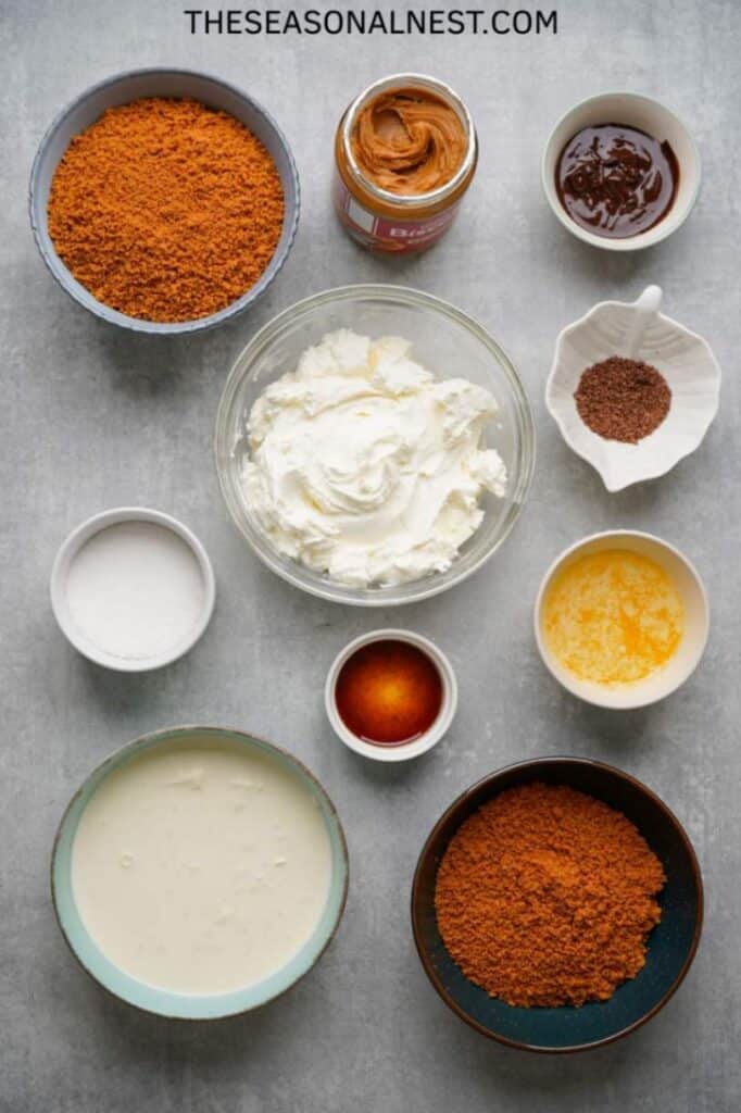 Flat lay of Biscoff cheesecake ingredients including cream cheese, Biscoff spread, whipped cream, cookie crumbs, sugar, butter, and vanilla.