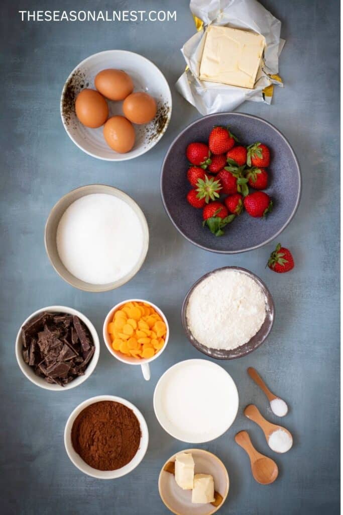 Flat lay of brownie ingredients, including eggs, butter, sugar, flour, cocoa powder, chocolate, cream, orange candy melts, strawberries, and small wooden spoons with baking soda and salt.