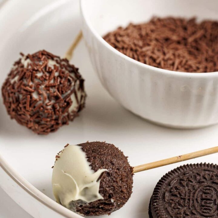 Oreo cookie cake pops on a plate with a bowl of chocolate sprinkles.