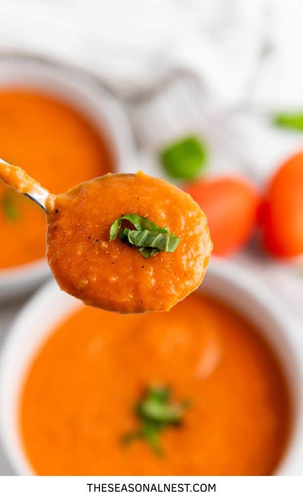 Close-up of a spoonful of roasted tomato basil soup topped with fresh basil, with bowls of soup in the background.