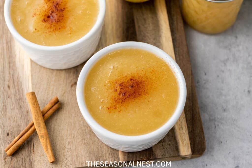 Two small white bowls of homemade Instant Pot applesauce topped with cinnamon, served on a wooden board with cinnamon sticks.