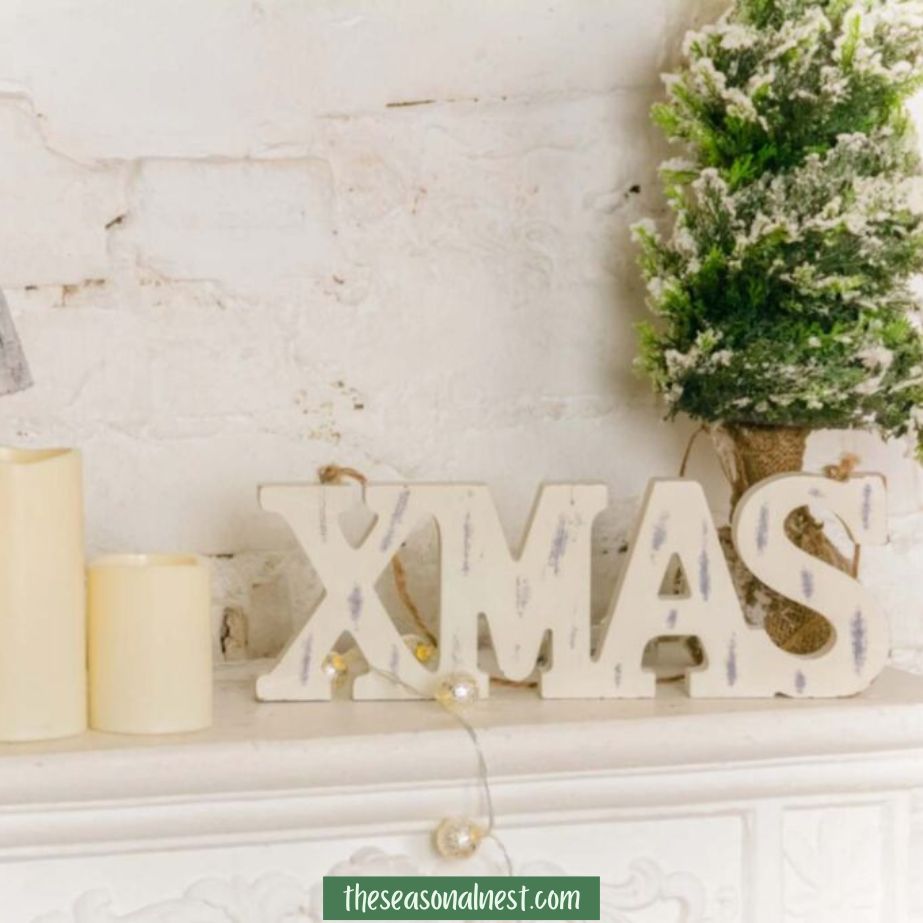 Wooden "XMAS" sign on a mantel with candles and a frosted tree.