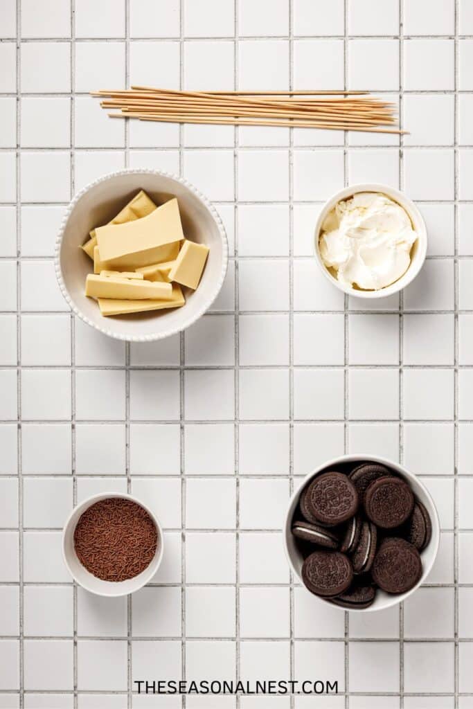 Ingredients for Oreo cake pops displayed on a tiled surface, including Oreos, cream cheese, white chocolate, sprinkles, and wooden sticks.