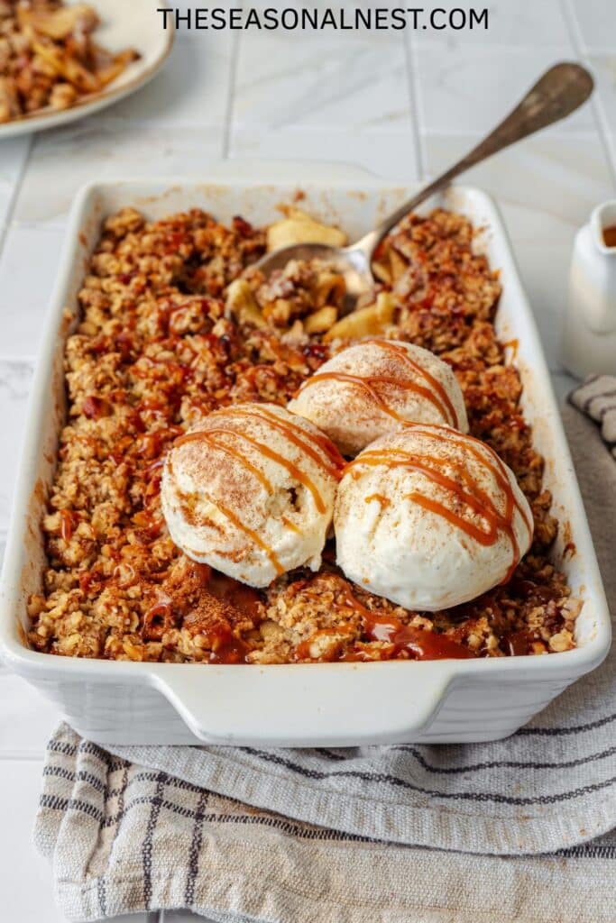 Baked apple crisp in a white pan topped with scoops of vanilla ice cream and caramel drizzle.