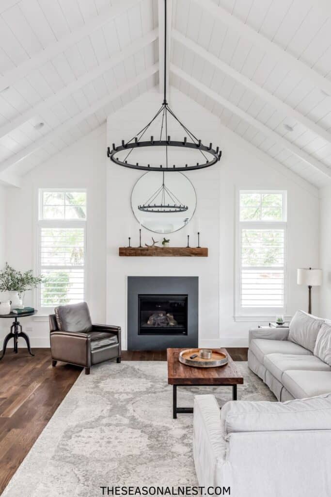 Bright white farmhouse living room with vaulted ceiling, black iron chandelier, leather accent chair, neutral sofas, and wood mantel over fireplace.