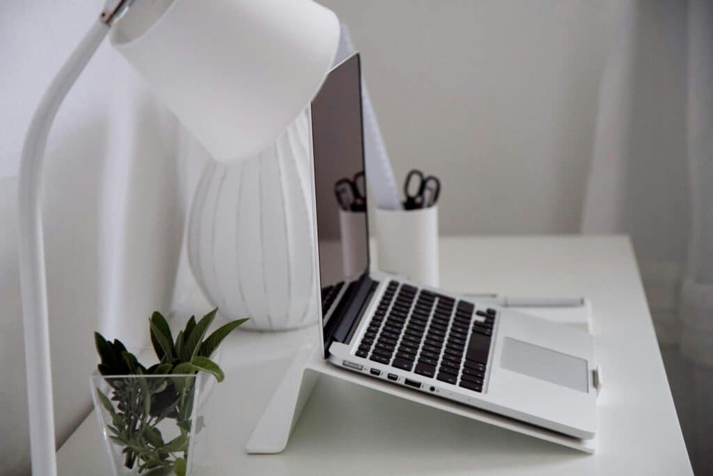 A clean home office study with a laptop, a lamp, scissors, a white vase and a planter.