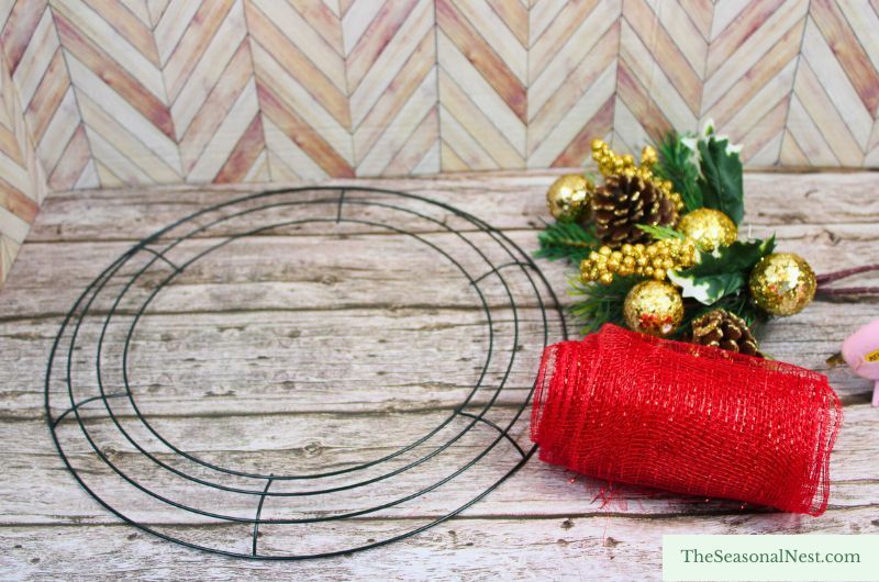 DIY Christmas wreath materials, including red deco mesh, gold pinecone picks, and a wire frame laid out on a rustic wooden surface