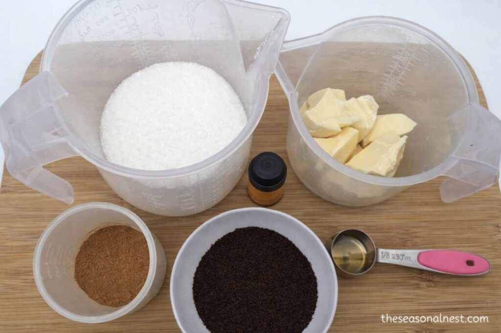 Top view of ingredients for homemade coffee scrub cubes including white sugar, cocoa butter chunks, ground coffee, cinnamon, essential oil bottle, and measuring spoon on a wooden board.