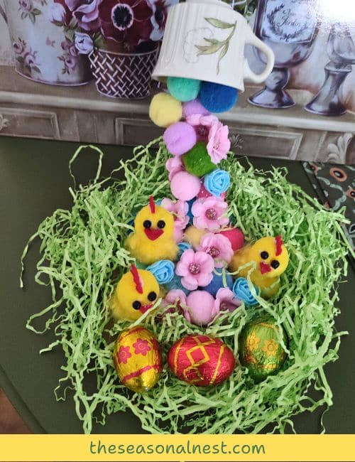 A DIY floating tea cup with pastel pom-poms, mini yellow chicks, and Easter eggs, resting on green shredded paper for a charming holiday display.