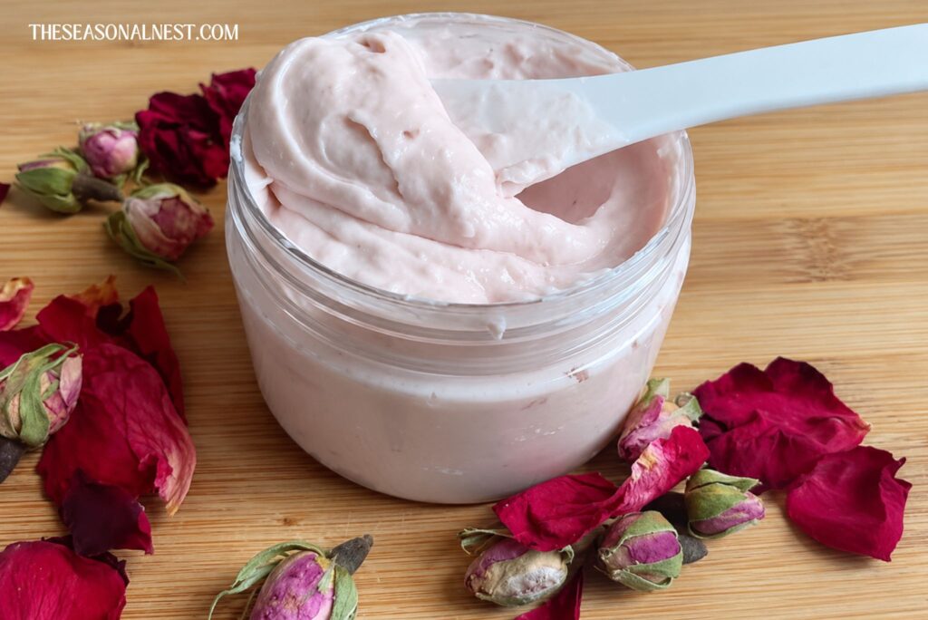 Homemade rose water face cream in a container with a spatula and scattered roses.