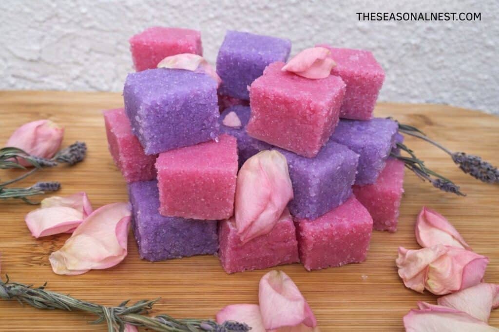 DIY sugar scrub cubes in lavender and rose colors with scattered rose petals and lavender sprigs.
