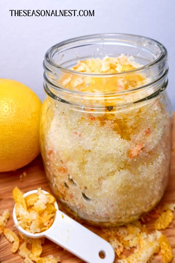 A lemon and DIY lemon sugar scrub in a glass jar with lemon peels in a spoon.