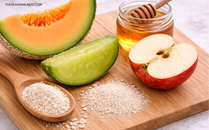 Fresh cantaloupe wedge, halved red apple, jar of honey with dipper, and rolled oats arranged on a wooden cutting board for a natural DIY face mask recipe.