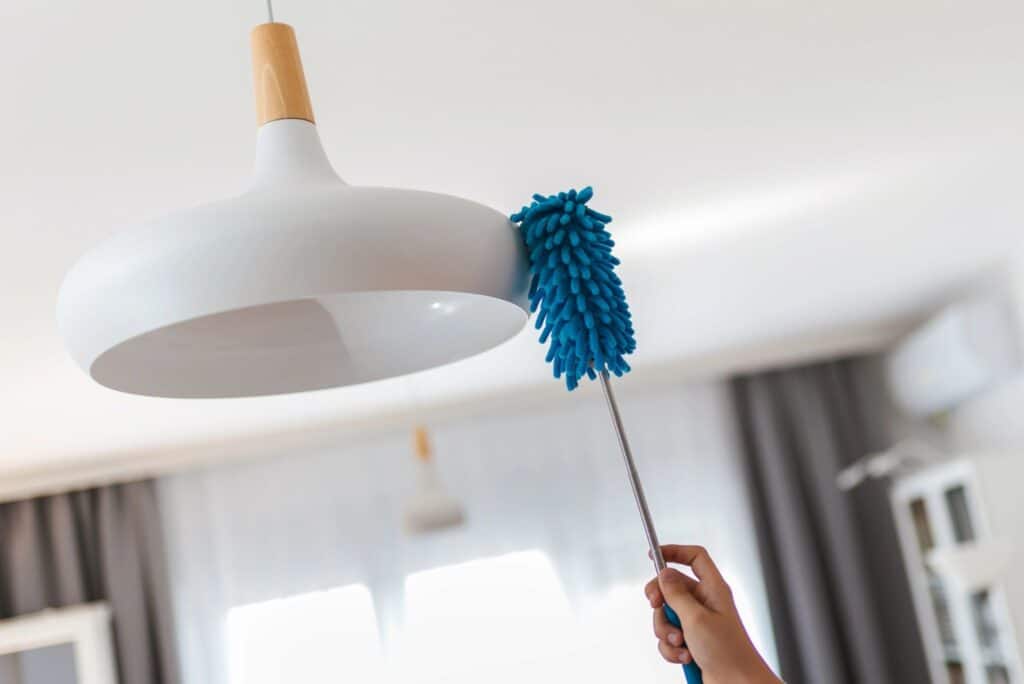 Hand using a blue microfiber duster to clean a white ceiling light fixture during spring cleaning.