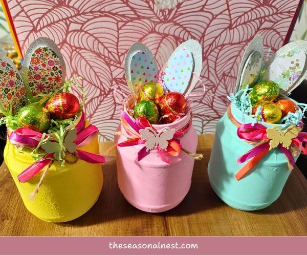 Three pastel-painted Easter mason jars with paper bunny ears, twine, satin ribbon, and wooden butterfly embellishments, filled with chocolates