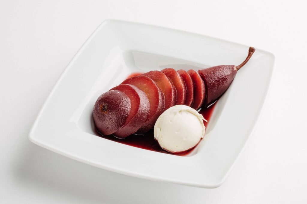 A red wine poached pear cut in slices with a scoop of vanilla ice cream