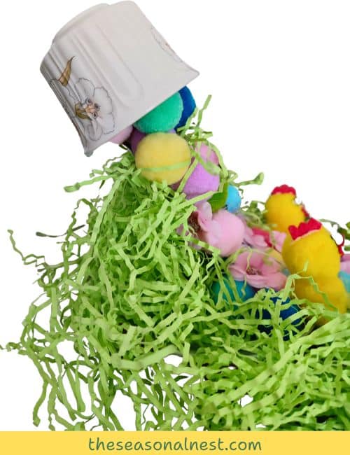 DIY floating tea cup decorated with pastel pom-poms, flowers, and small yellow chicks, creating a charming Easter centerpiece with green shredded paper and decorative eggs.