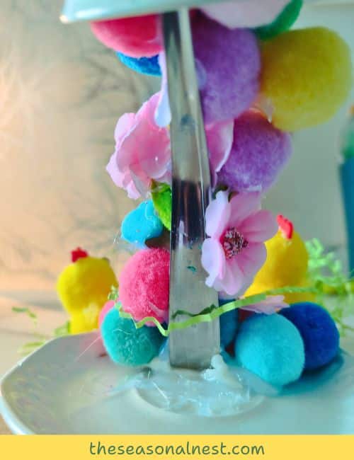 A spoon is secured to a saucer with hot glue, decorated with colorful pom-poms, tiny flowers, and chicks to create an Easter floating tea cup topiary.