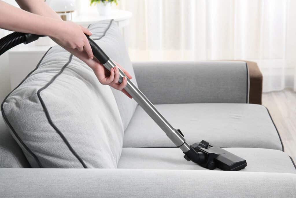 Vacuum cleaner nozzle cleaning a grey fabric sofa cushion during spring deep cleaning.