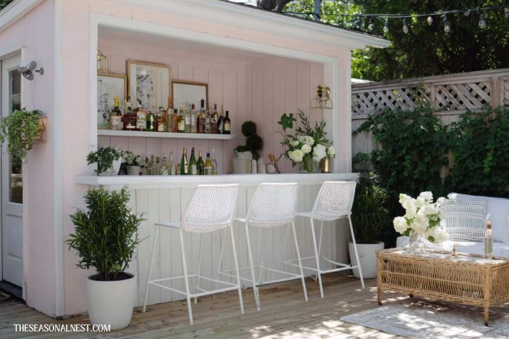 Soft pink outdoor bar with white stools, shelves, bottles, plants, and seating area in a bright patio setting.