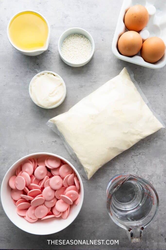 Ingredients for pink cake pops laid out on a counter, including cake mix, eggs, oil, frosting, pink candy melts, sprinkles and water.