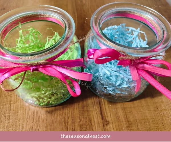 Two small Easter mason jars filled with pastel shredded paper, tied with twine and pink ribbon.