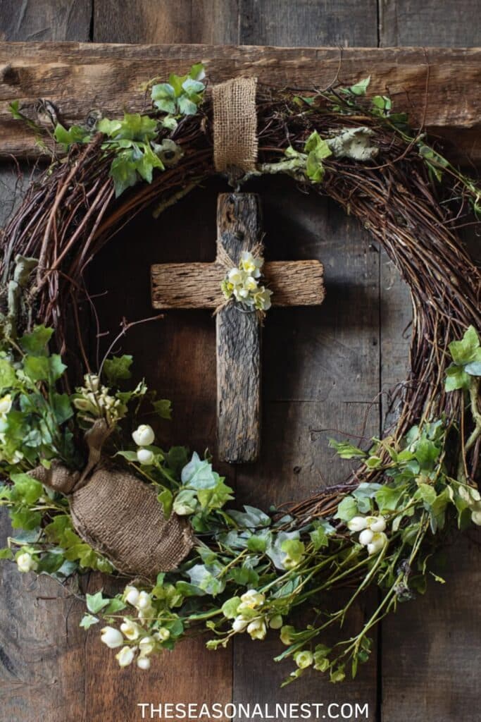 Rustic grapevine front door wreath with a hanging wooden cross, ivy greenery, white spring flowers, and burlap accents on a dark wood door.