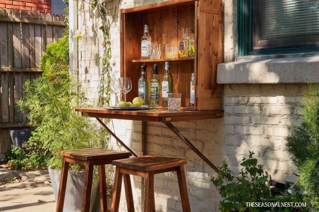 Wall mounted wooden drinks station with fold down table, shelves, bottles, and stools in a small patio setting.