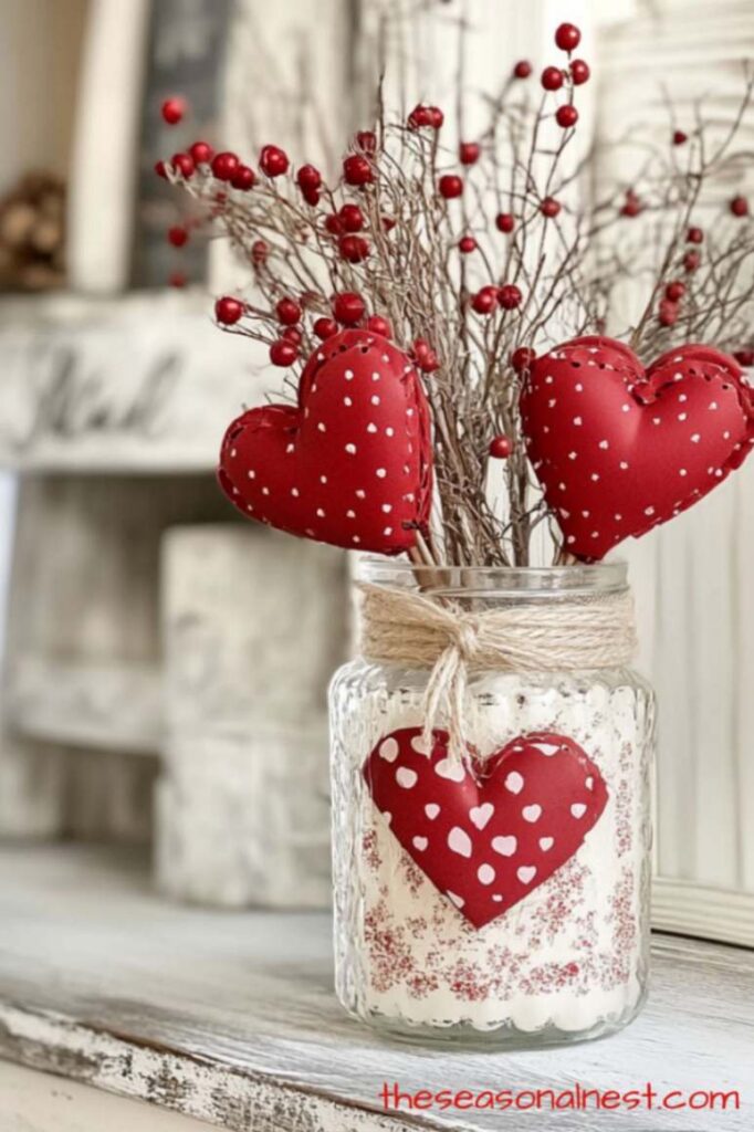 Valentine mason jar centerpiece with red heart decor, twine wrapped jar, white painted finish, and berry branches on a rustic table.